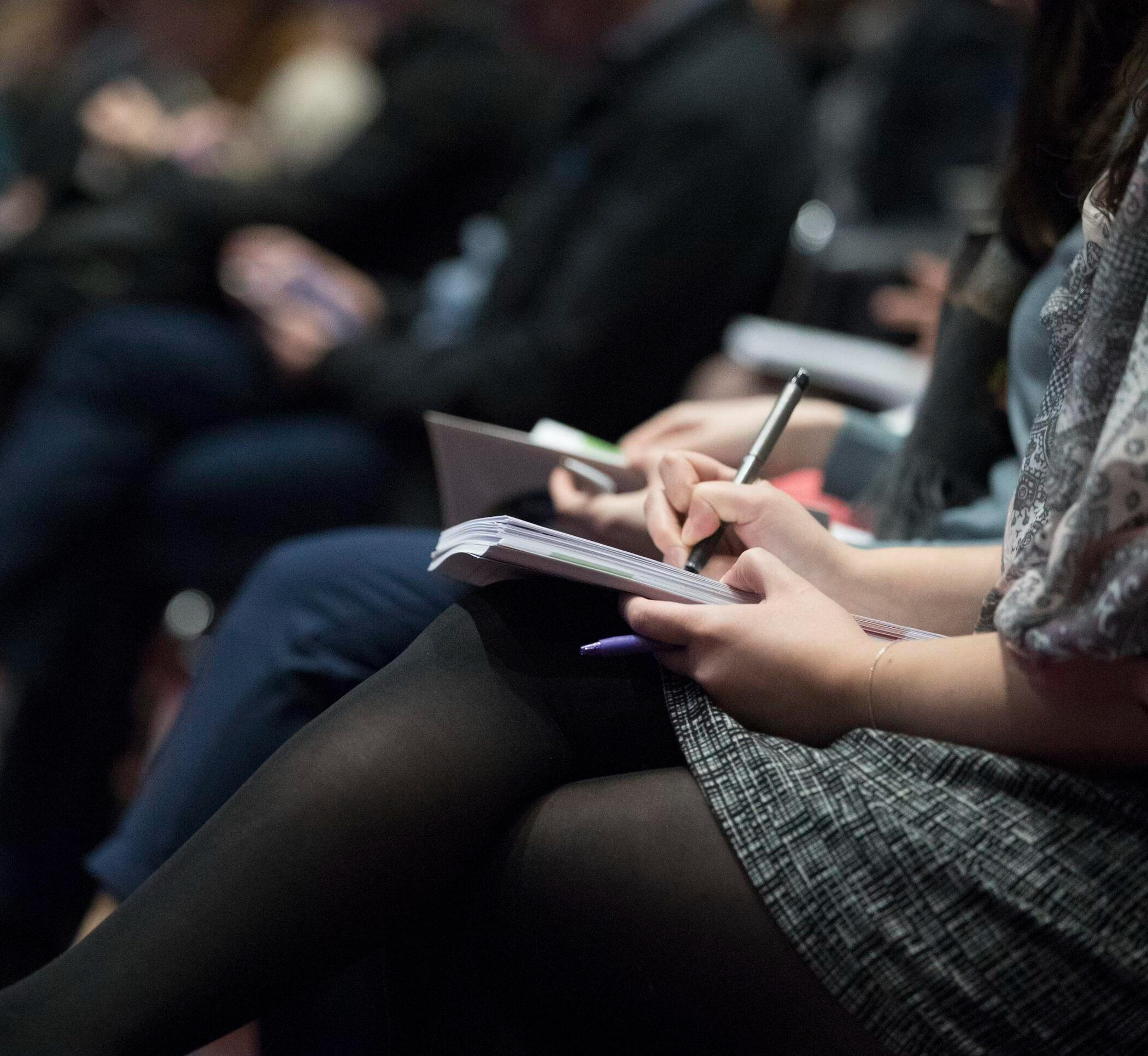 Leadership Seminar – Participants Taking Notes Close-up of professionals seated at a seminar or conference, focused on a woman writing notes in a notebook. The scene captures active participation and engagement during a business or leadership event.