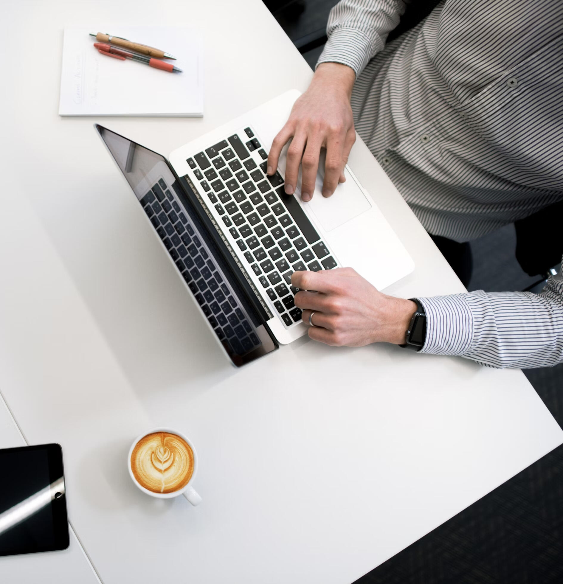 Executive Workspace Overhead Top-down view of a business professional working on a laptop at a clean white desk, with a notepad, coffee, conference phone, remote control, and tablet in view.